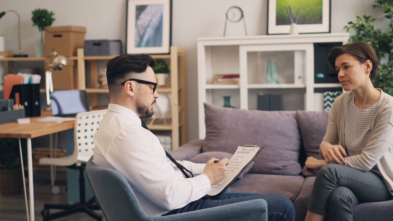 hero-img A therapist engages in a consultation session with a female patient in a cozy office setting.