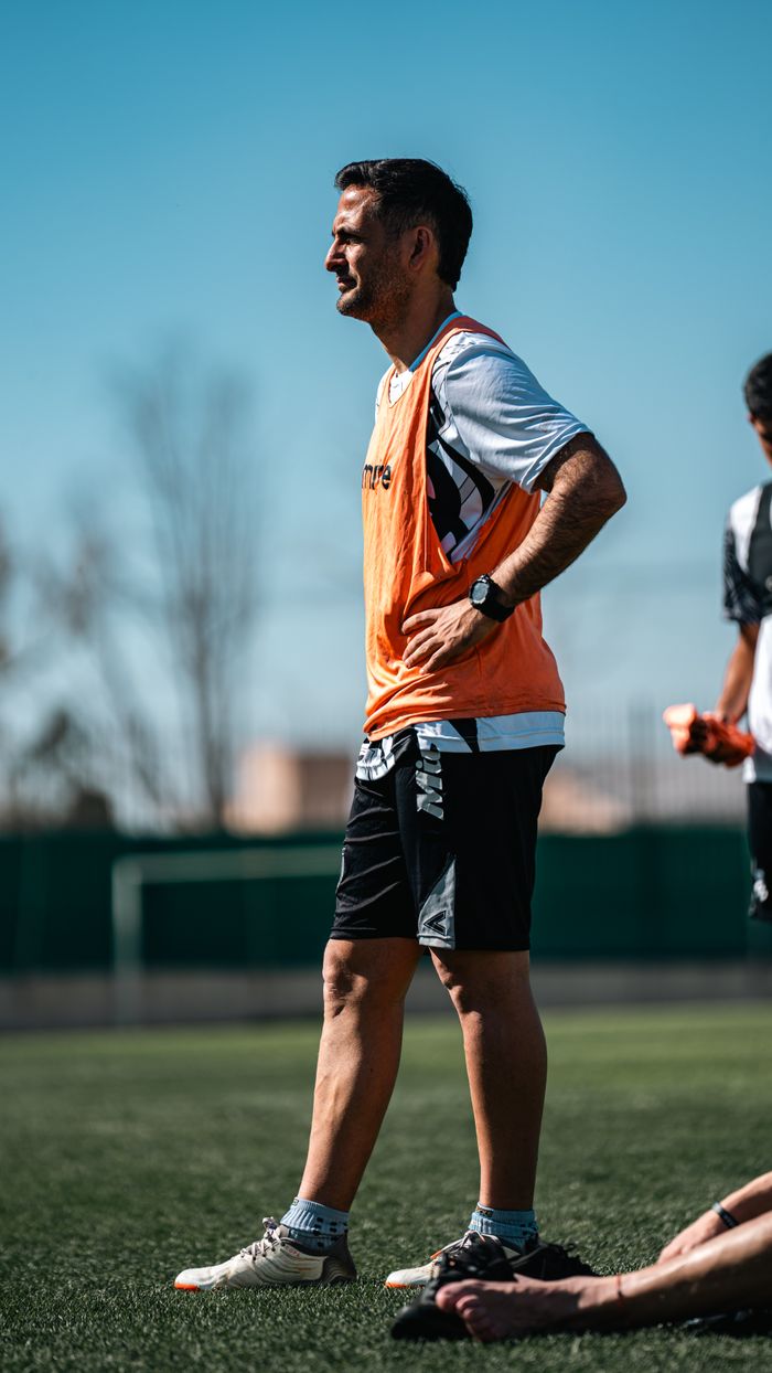 A soccer coach in an orange vest instructing players on an outdoor field during the day.