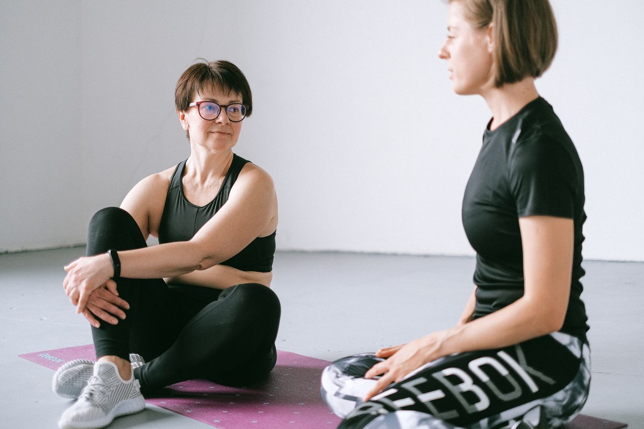 service-01 Two women in activewear discuss fitness on a yoga mat indoors.